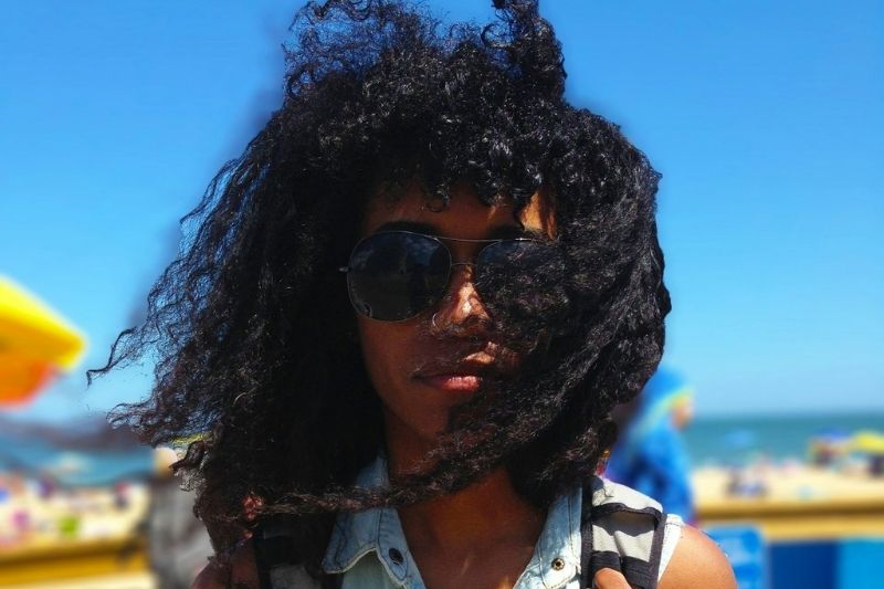 Femme aux cheveux bouclés au vent sur une plage en été, illustrant les effets de la chaleur et du vent sur les cheveux secs.